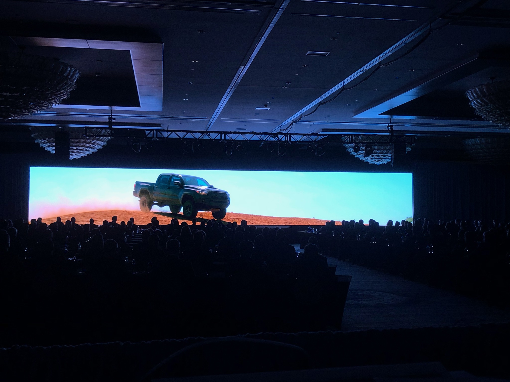 Audience watches a video of a Toyota truck driving off-road on a wide LED screen during a Gulf States Toyota event in a dark ballroom setting.