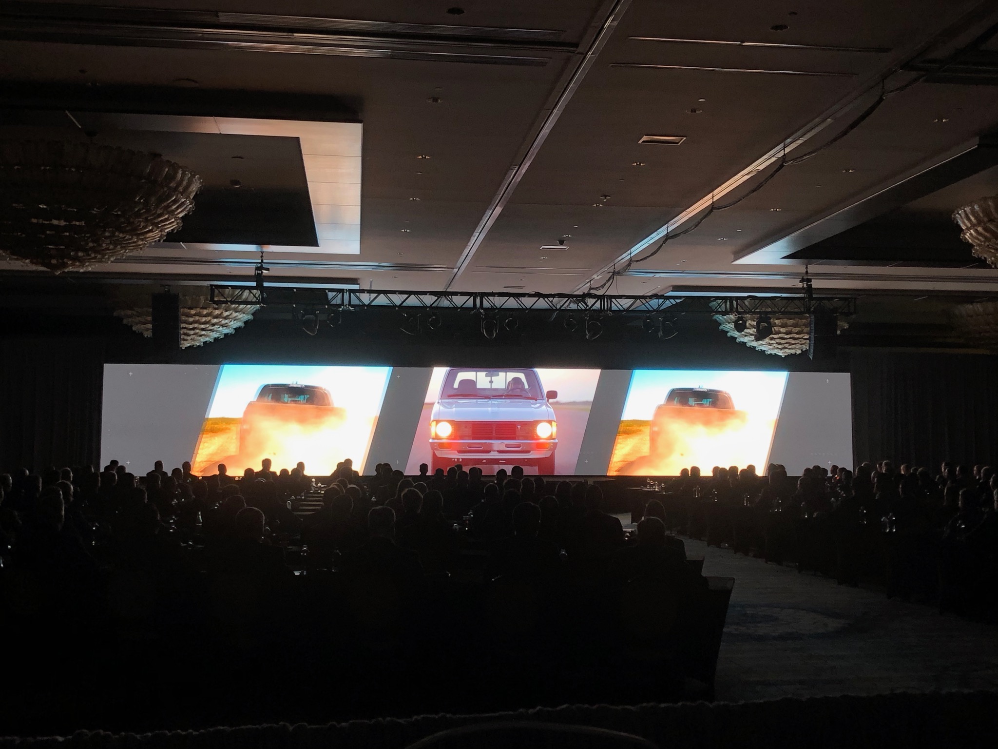 Attendees watch a wide-screen video of trucks driving through dust during a Gulf States Toyota event in a dark, chandelier-lit ballroom.