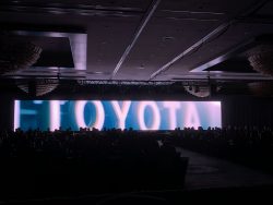 Audience watches a widescreen video displaying the word “TOYOTA” during a Gulf States Toyota event in a dimly lit ballroom with chandeliers.