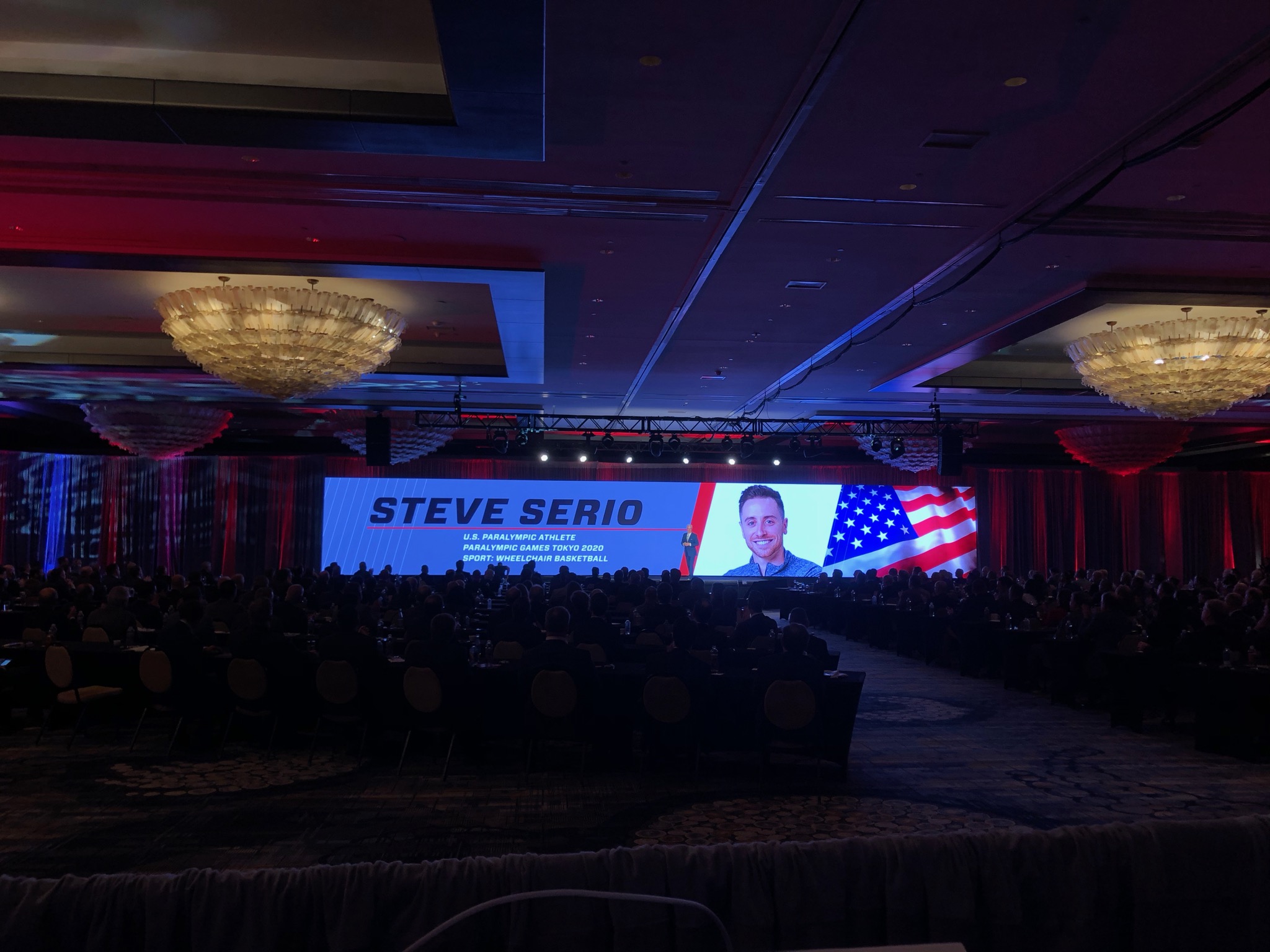 Steve Serio, U.S. Paralympic wheelchair basketball athlete, speaks at a Gulf States Toyota event with his name and photo on a large LED screen.