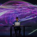 Man sitting at desk in front of computer with 3D projection of car in background