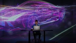 Man sitting at desk in front of computer with 3D projection of car in background