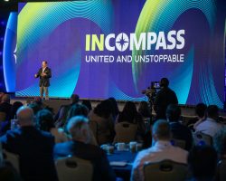 A speaker presents on stage at a corporate event with "INCOMPASS: UNITED AND UNSTOPPABLE" displayed on a large screen behind him.