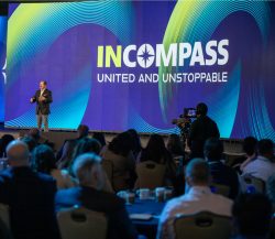 A speaker presents on stage at a corporate event with "INCOMPASS: UNITED AND UNSTOPPABLE" displayed on a large screen behind him.
