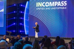 A speaker presents at the INCOMPASS event, with "UNITED AND UNSTOPPABLE" displayed on a large screen behind him to an engaged audience.
