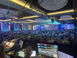 A large audience attends the INCOMPASS event, viewed from the production control area with screens, soundboards, and stage lighting setup.