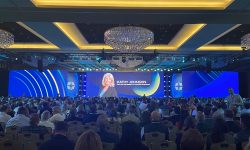 A large audience watches a corporate event with "Kathy Johnson, Executive VP of Strategic Development" displayed on stage screens.