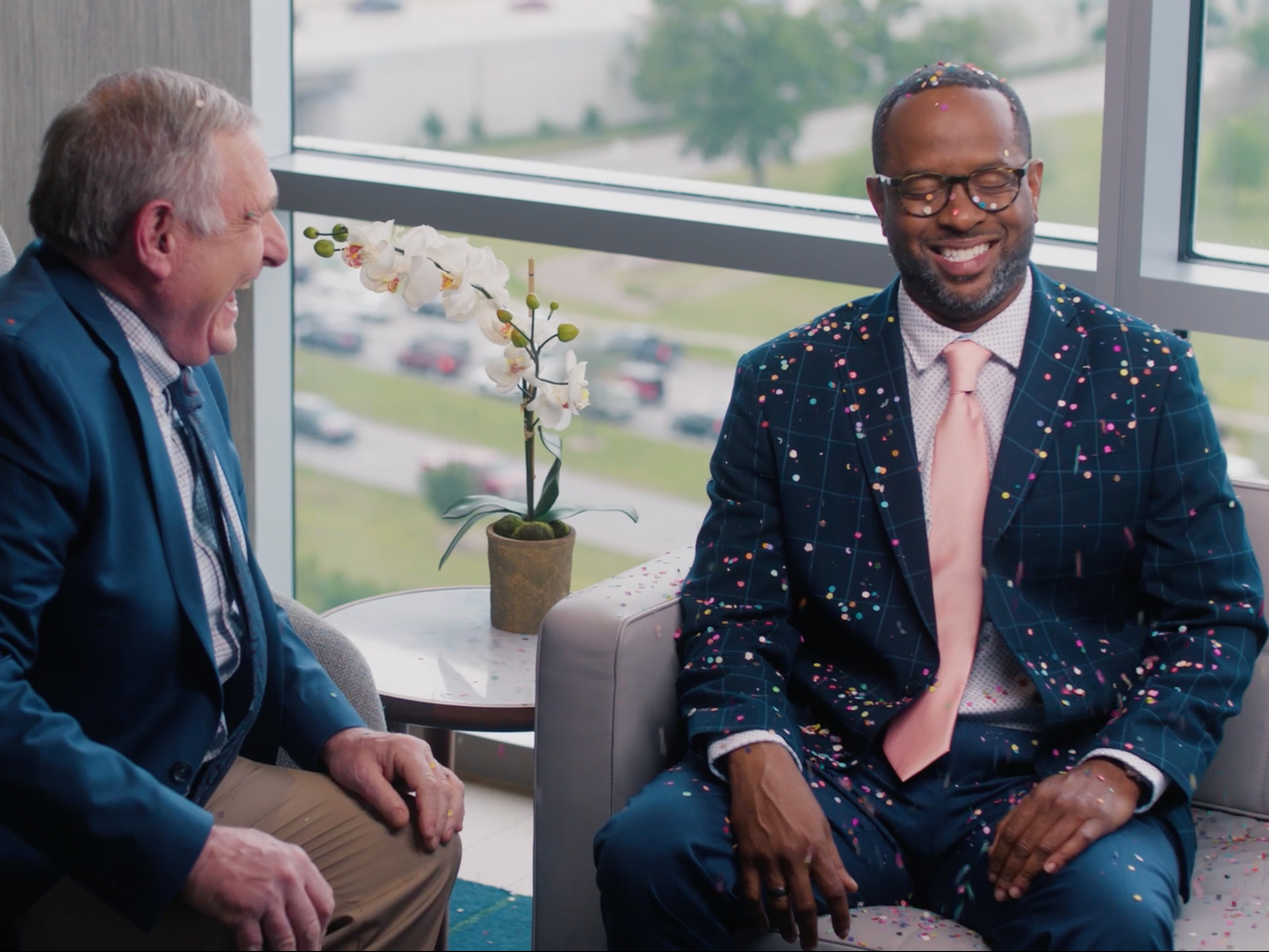 Two men in suits laugh, one covered in colorful confetti, sitting in an office with a large window and an orchid on the table.