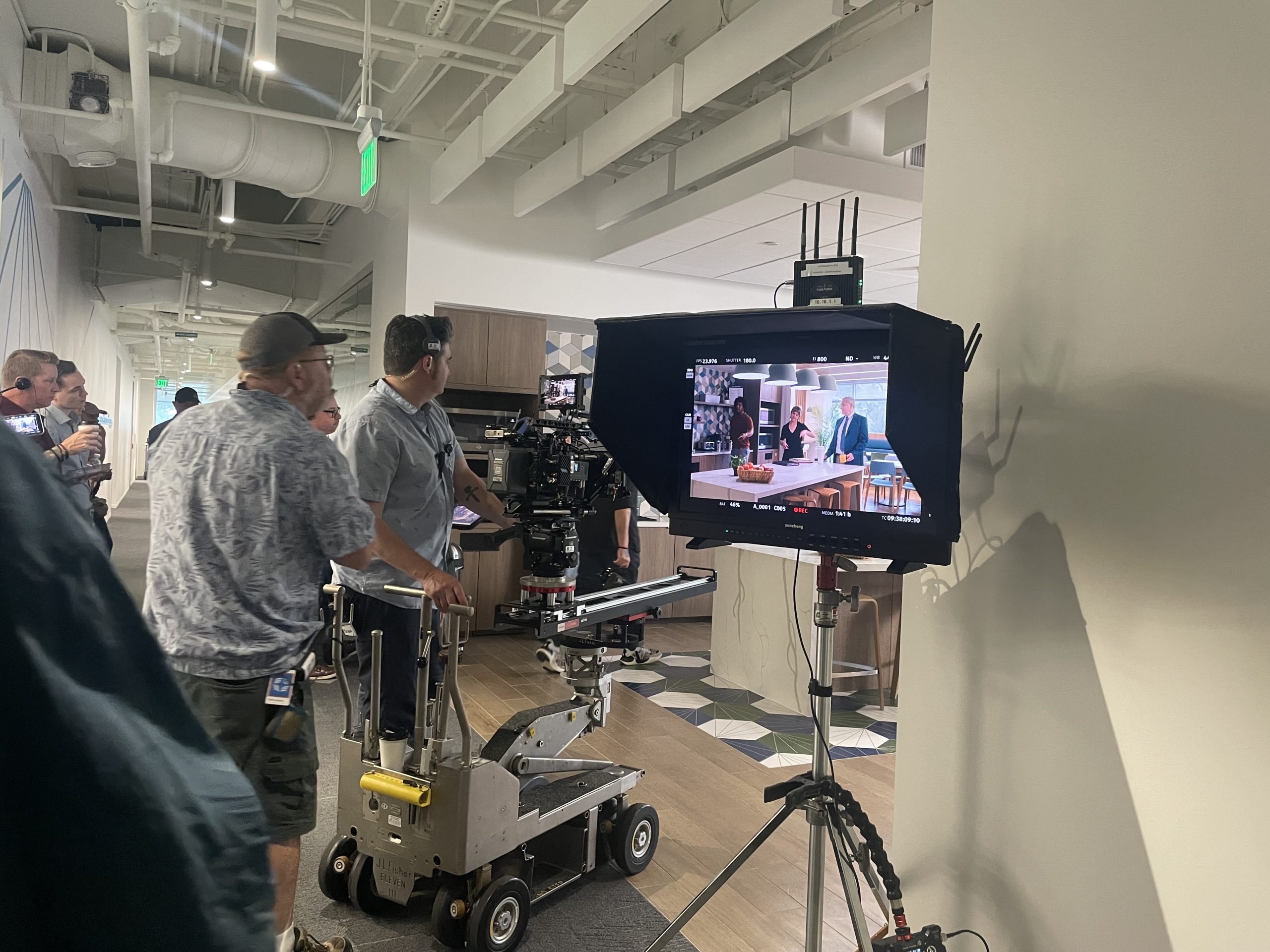 A film crew operates a camera on a dolly, capturing a scene displayed on a monitor in a modern office hallway.