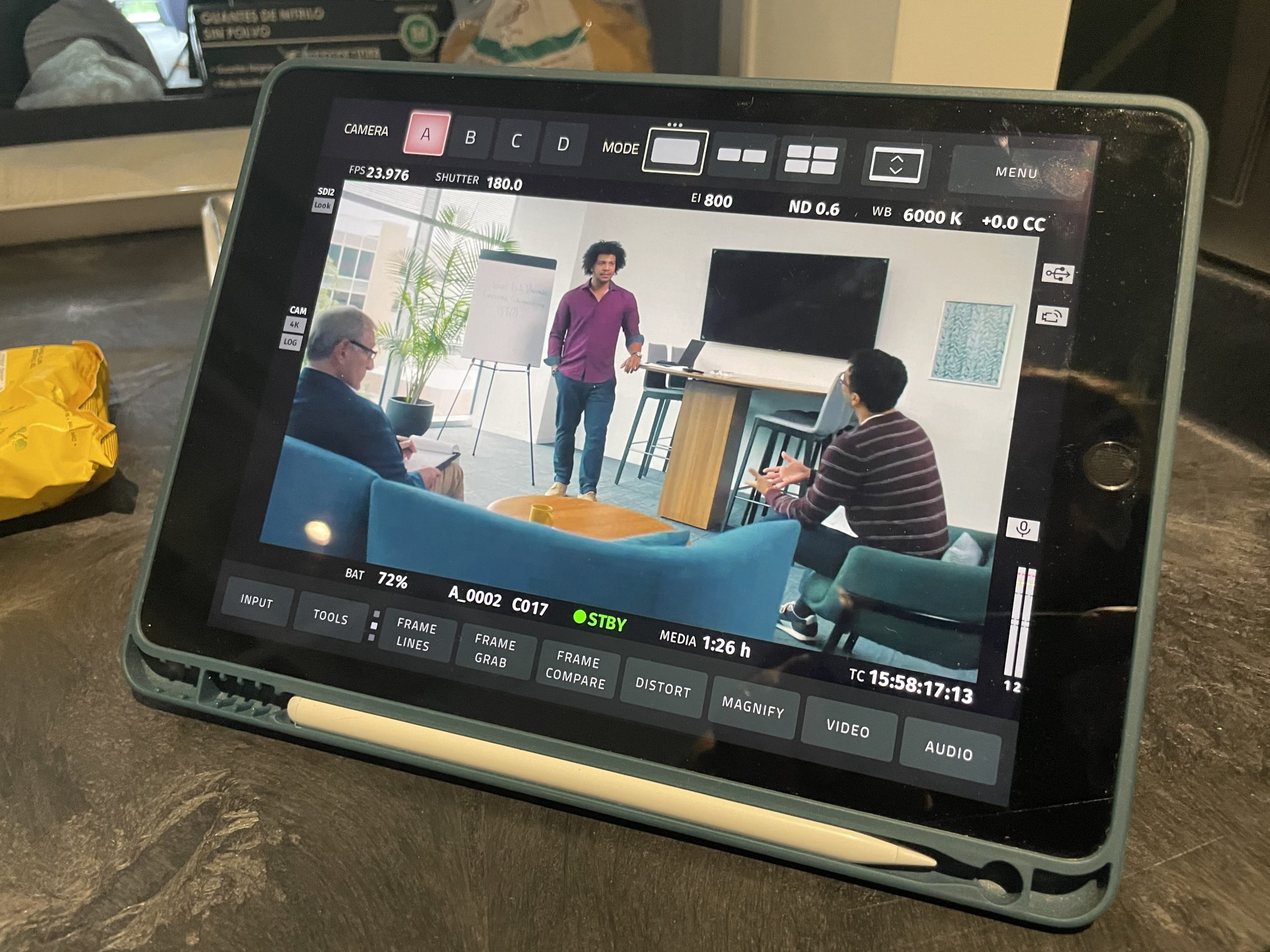 A tablet displaying a video shoot setup with a man presenting to two seated individuals, viewed through a camera monitor.