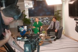 Behind-the-scenes of a video shoot: two people seated and talking on set while a cameraman captures the scene.