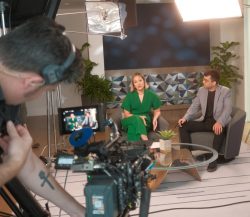 Behind-the-scenes of a video shoot: two people seated and talking on set while a cameraman captures the scene.