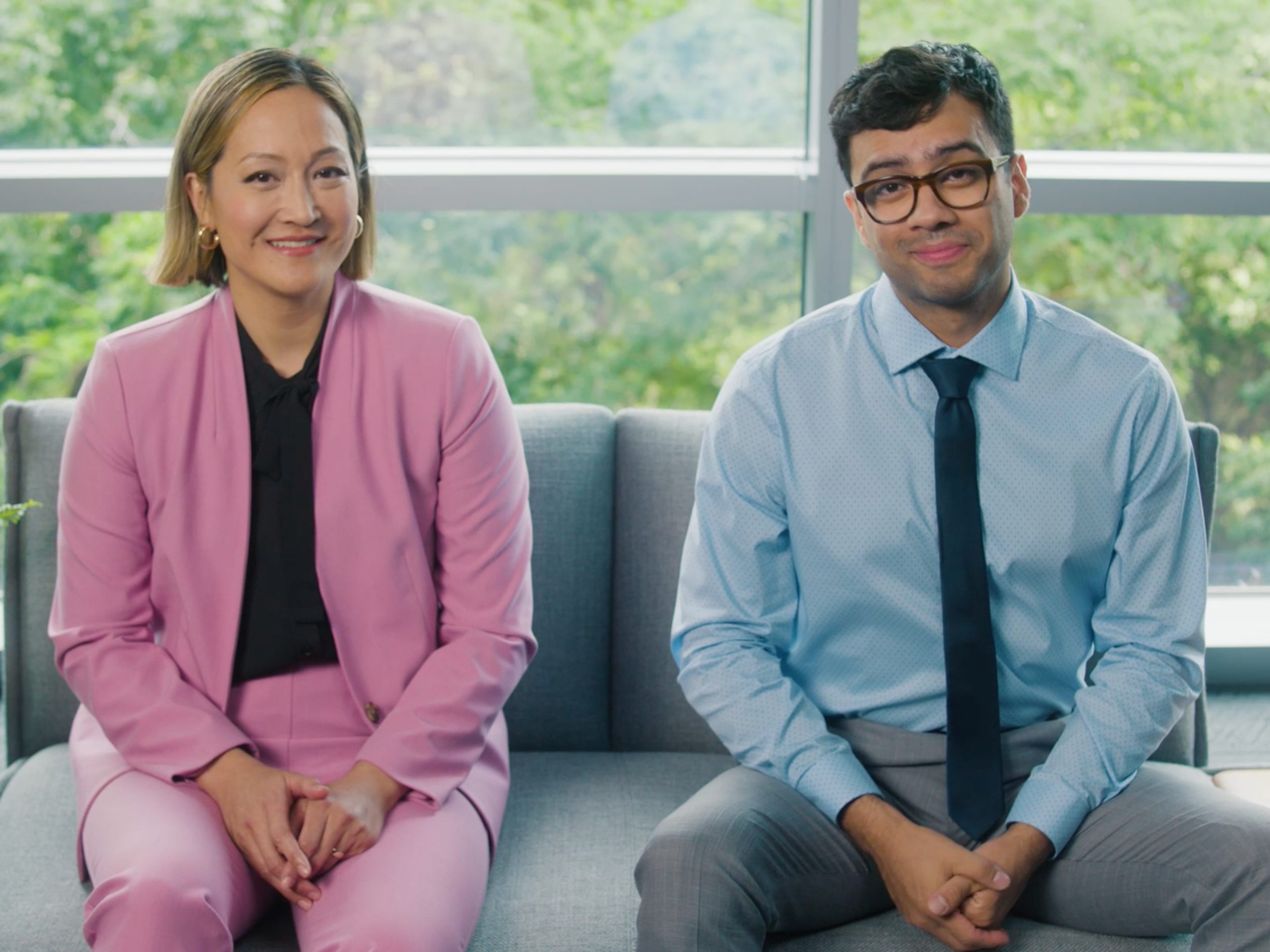 Two professionals sitting on a couch, smiling, in a well-lit office setting with large windows and greenery in the background.