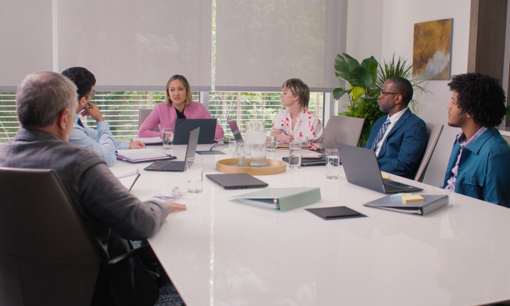 A diverse team sits in a bright conference room engaged in a meeting, with laptops, notebooks, and water on the table.