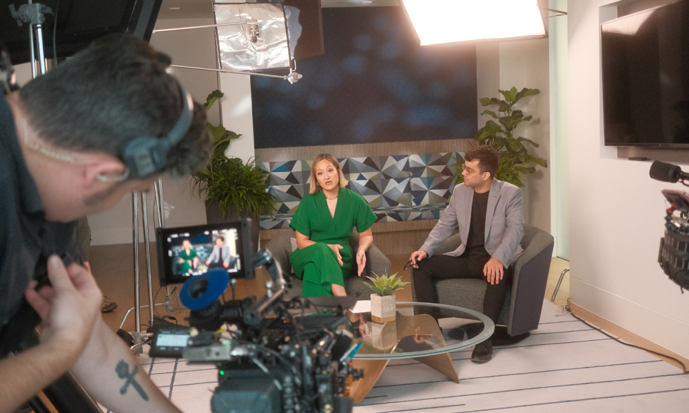 Behind-the-scenes of a video shoot: two people seated and talking on set while a cameraman captures the scene.