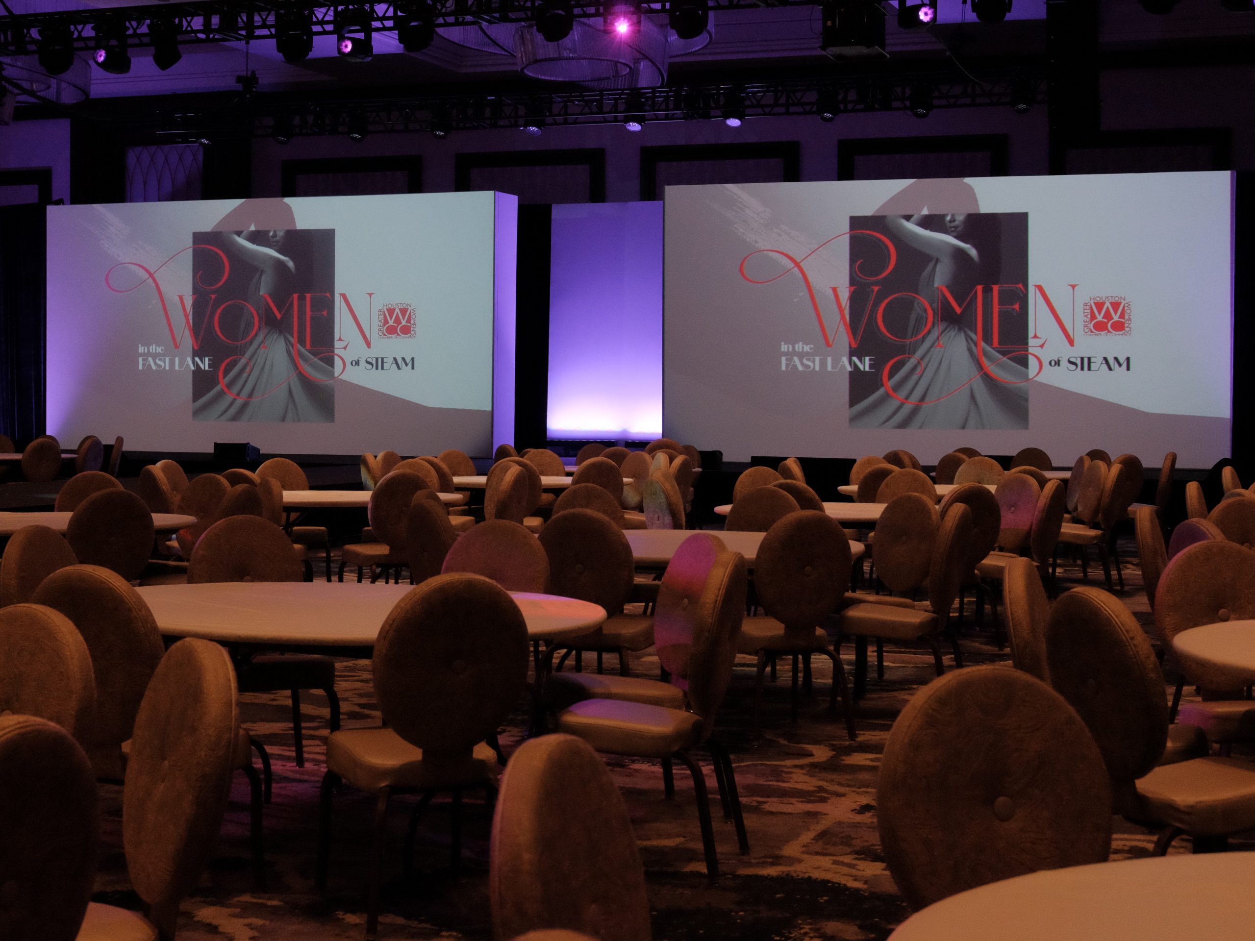 Event setup for Women in the Fast Lane of STEAM with large screens displaying the event logo, surrounded by round tables and elegant seating.