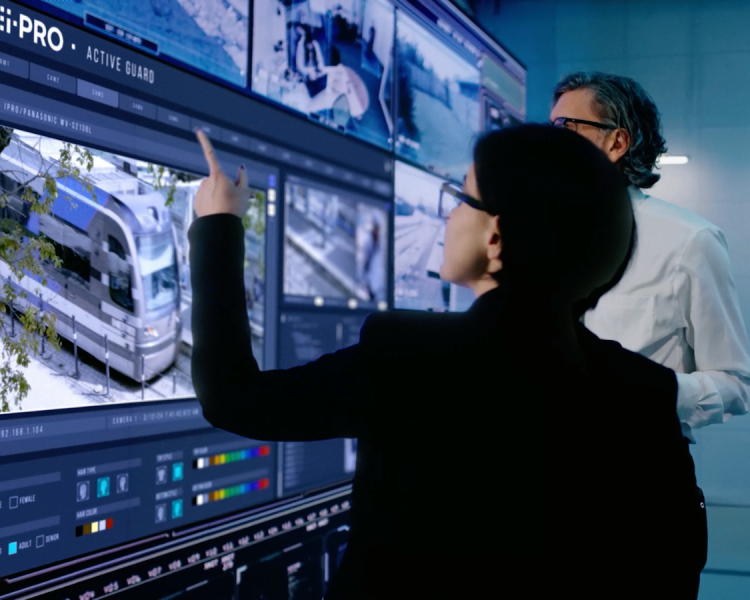 Two security professionals analyze surveillance footage on a large screen displaying i-PRO’s Active Guard system, which monitors multiple feeds and data.