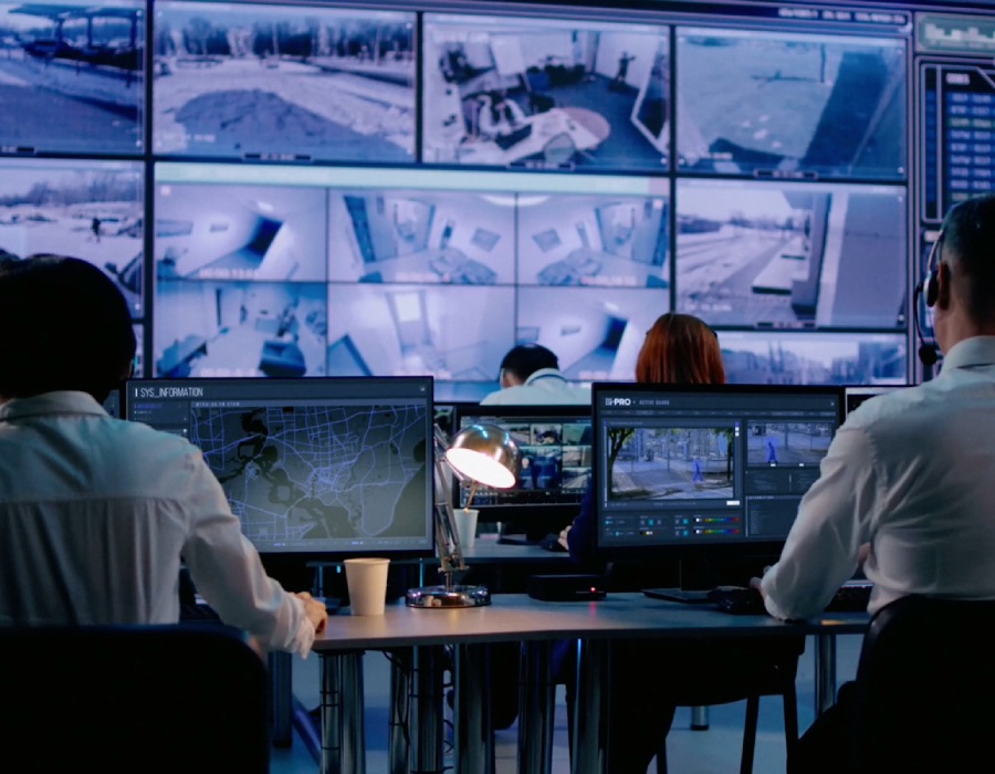 A group of security operators monitor multiple surveillance feeds on large and small screens in a control room, using i-PRO’s security system.