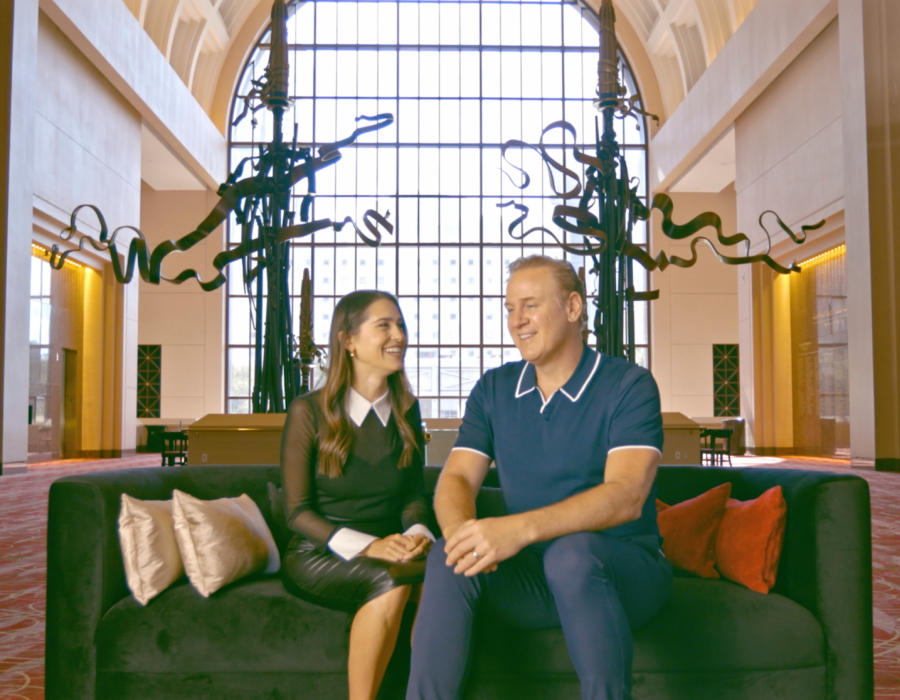 A smiling couple sits on a black couch in a grand hall with large windows and abstract black sculptures, engaged in conversation.