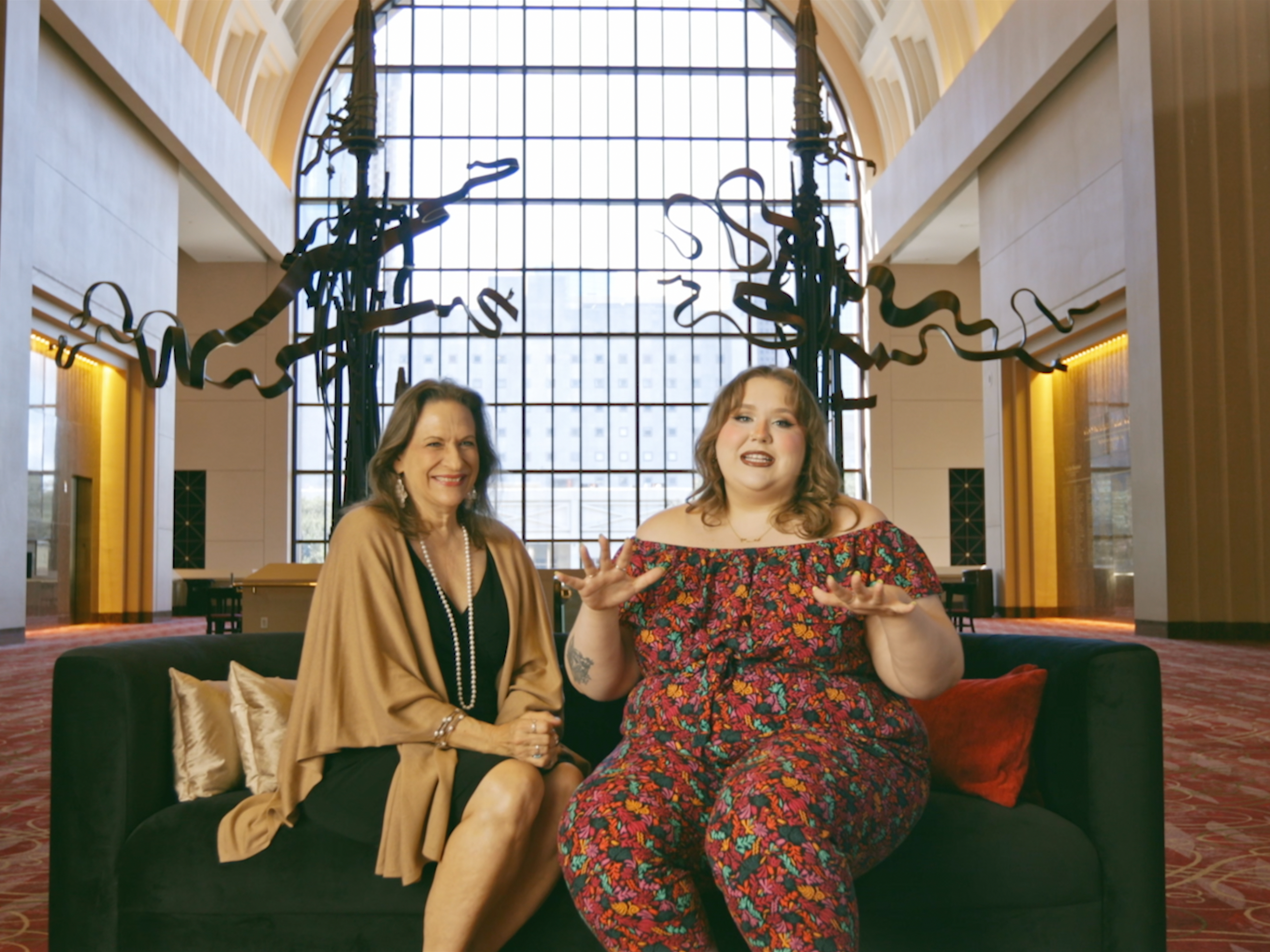 Two women sit on a black couch in a grand hall, engaged in conversation. One gestures expressively while the other smiles warmly.