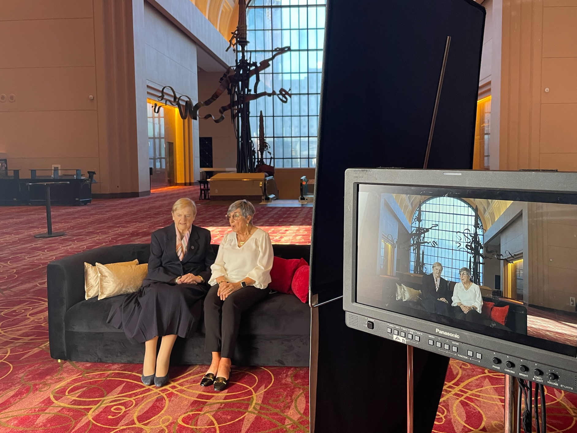 Two women sit on a black couch in a grand hall during a video shoot. A monitor in the foreground displays their interview setup.