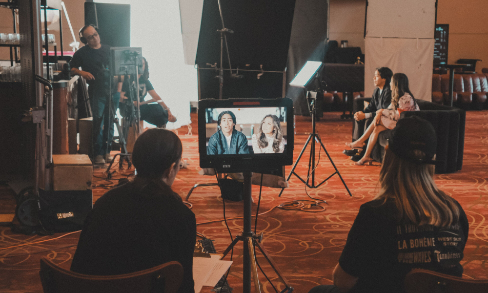 A behind-the-scenes look at a video shoot shows a monitor displaying two speakers, with crew members and lighting equipment in the background.