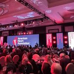 Guests seated at a gala event with American Heart Association branding on a large LED stage display and red lighting throughout the venue.