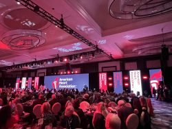 Guests seated at a gala event with American Heart Association branding on a large LED stage display and red lighting throughout the venue.